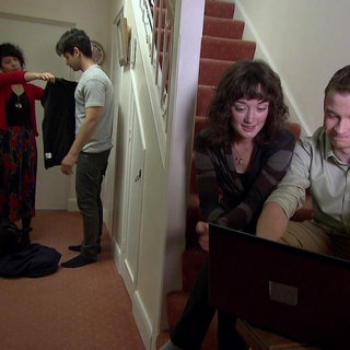 Ein junger Mann und eine junge Frau sitzen auf einer Treppe und schauen auf einen Computer. Neben der Treppe probieren zwei andere junge Erwachsene Kleidung an.