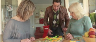 Drei Personen und eine rote Strumpfhandpuppe sind an einem Tisch versammelt und spielen ein Spiel.