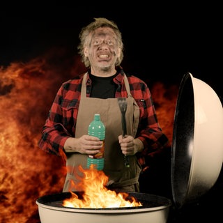 Ein Mann mit rußgeschwärztem Gesicht und Spiritusflasche in der Hand steht hinter einem Grill, aus dem eine Stichflamme schlägt.