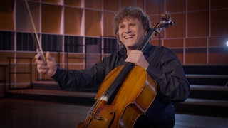 Ein Mann beendet freudestrahlend ein Musikstück auf einem Violoncello.  Das Instrument gehört zur Gruppe der Streicher.