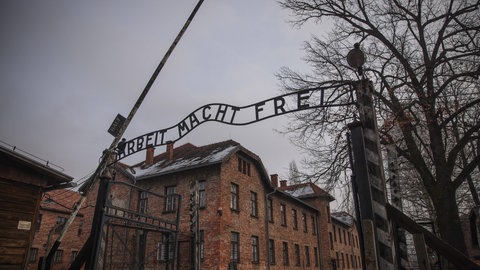 Der Eingang zum Konzentrationslager Auschwitz mit dem Schriftzug „Arbeit macht frei“. Dahinter mehrstöckige Backsteingebäude und rechts Bäume. Mit dem Schriftzug haben die Nazis die Häftlinge verhöhnt.
