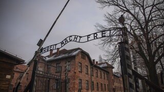 Der Eingang zum Konzentrationslager Auschwitz mit dem Schriftzug „Arbeit macht frei“. Dahinter mehrstöckige Backsteingebäude und rechts Bäume. Mit dem Schriftzug haben die Nazis die Häftlinge verhöhnt.