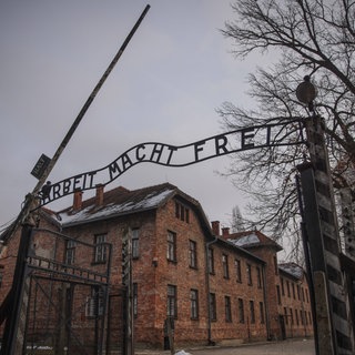 Der Eingang zum Konzentrationslager Auschwitz mit dem Schriftzug „Arbeit macht frei“. Dahinter mehrstöckige Backsteingebäude und rechts Bäume. Mit dem Schriftzug haben die Nazis die Häftlinge verhöhnt.