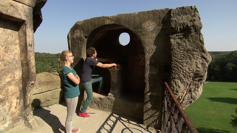 Ein Mann und eine Frau stehen auf einer Steinmauer und gucken durch ein Felsloch über grüne Felder.