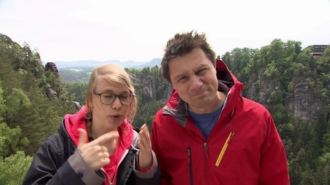 Eine Frau und ein Mann stehen in einem Gebirge, umgeben von Wäldern.