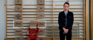 Ein junger Mann steht vor einer Sprossenwand, an ihr hängen Schilder. Darauf stehen französische Pronomen und die Endungen konjugierter Verben auf -er.