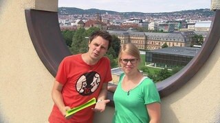 Ein Mann und eine Frau stehen vor einem runden Loch in einer Mauer, im Hintergrund eine Stadt.