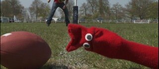 Eine rote Strumpfhandpuppe liegt auf dem Rasen, neben ihr ein Rugbyball.