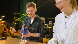 Ein Mann und eine Frau im Laborkittel mit Schutzbrillen richten die Flamme einer Lötlampe auf Plastikteile in einem Schmelztiegel