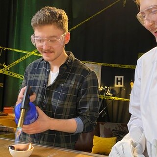 Ein Mann und eine Frau im Laborkittel mit Schutzbrillen richten die Flamme einer Lötlampe auf Plastikteile in einem Schmelztiegel