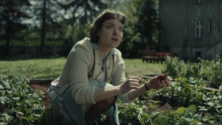 Spielszene: Hedwig Höß hockt im Erdbeerbeet im Garten neben dem KZ Auschwitz. Sie schaut zum Himmel, von dem es Asche aus dem angrenzenden KZ regnet.