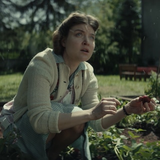 Spielszene: Hedwig Höß hockt im Erdbeerbeet im Garten neben dem KZ Auschwitz. Sie schaut zum Himmel, von dem es Asche aus dem angrenzenden KZ regnet.