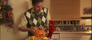 Ein Mann und eine rote Strumpfhandpuppe höhlen einen Kürbis für Halloween aus.