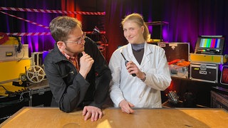 Ein Mann und eine Frau in einem Fernsehstudio. Die Frau trägt einen Laborkittel und hält ein brennendes Gasfeuerzeug in der Hand