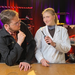 Ein Mann und eine Frau in einem Fernsehstudio. Die Frau trägt einen Laborkittel und hält ein brennendes Gasfeuerzeug in der Hand