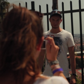 Ein junger Mann steht vor einem Zaun, hinter ihm liegt die Stadt. Eine Frau macht ein Foto von ihm.