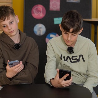 Zwei Schüler sitzen in einem modernen Klassenzimmer. Beide schauen genervt auf ihr Smartphone.