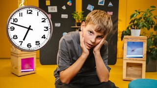 Schüler sitzt genervt in einem modernen Klassenzimmer, an der Wand hängt eine große Uhr