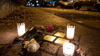 Stolpersteine im Boden, daneben Kerzen und weiße Rosen. Es ist Nacht. Die Stolpersteine erinnern an jüdische Menschen, die von den Nazis verfolgt und getötet wurden.