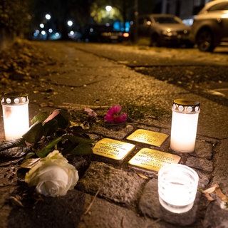 Stolpersteine im Boden, daneben Kerzen und weiße Rosen. Es ist Nacht. Die Stolpersteine erinnern an jüdische Menschen, die von den Nazis verfolgt und getötet wurden.