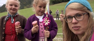 Eine Frau und zwei junge Mädchen in bayrischer Tracht stehen vor grünen Wiesen und Wäldern.