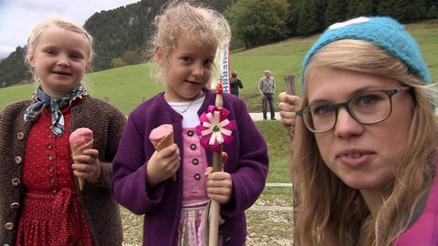 Eine Frau und zwei junge Mädchen in bayrischer Tracht stehen vor grünen Wiesen und Wäldern.