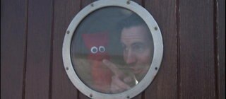 Ein Mann und eine rote Strumpfhandpuppe sind auf einem Schiff und schauen durch ein Bullauge.