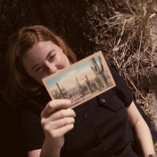 Spielszene: Joan Hinton hält lächelnd eine Postkarte in der Hand. Die Kernphysikerin arbeitet später am Manhattan-Projekt – der Entwicklung der Atombombe.