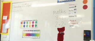 Eine Schultafel im Englischunterricht, davor eine rote Strumpfhandpuppe.