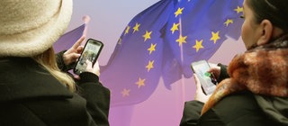 Zwei Jugendliche mit Handys vor EU-Flagge. Was ist die EU? Wie funktioniert die Zusammenarbeit in Politik und Wirtschaft in der Europäischen Union? 