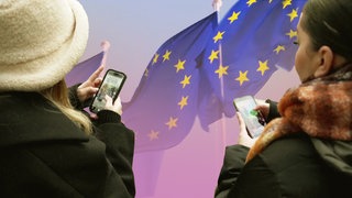 Zwei Jugendliche mit Handys vor EU-Flagge. Was ist die EU? Wie funktioniert die Zusammenarbeit in Politik und Wirtschaft in der Europäischen Union? 
